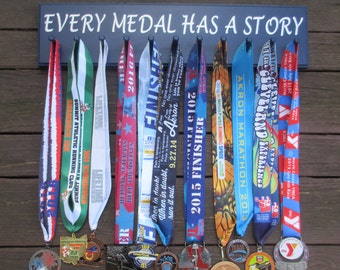 Könnte beinhalten: Ein schwarzes Schild mit weißem Text, der "Every Medal Has A Story" lautet, hängt an einer Holzwand. Neun Medaillen mit bunten Bändern sind an Haken unter dem Schild angebracht.
