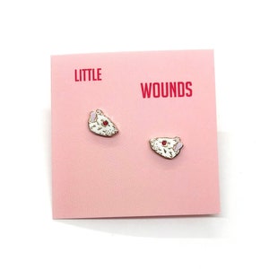 Op de afbeelding: Een paar witte emaille rattenkop oorbellen met roze accenten. De oorbellen zijn op een roze kaart met de tekst "LITTLE WOUNDS" in rood.