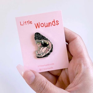 Peut inclure: Une épingle en émail noir représentant une illustration détaillée d'une gueule d'animal qui grogne avec des dents acérées. L'épingle est sur une carte rose avec le texte "Little Wounds" et l'adresse du site Web "littlewounds.com" et le nom du compte de médias sociaux "@littlewounds".
