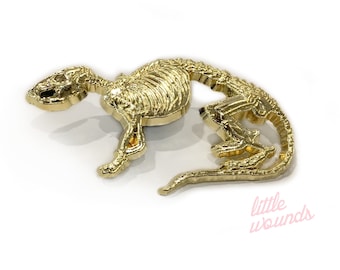 Op de afbeelding: Een gouden metalen pin met een skelet van een rat. De rat staat in een rennende houding met zijn staart opgerold. De pin is ongeveer 5 cm lang. De tekst "little wounds" is zichtbaar in de rechterbenedenhoek van de afbeelding.