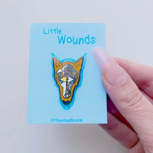 May include: A blue card with the text "Little Wounds" and a gold and multi-colored enamel pin of a fox skull. The website "littlewounds.com" is printed on the card.