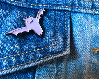 Puede incluir: Un broche de esmalte de color morado claro con forma de murciélago volando, con una cara sonriente sencilla y contornos negros, sujeto a una tela de mezclilla azul con costuras amarillas visibles a lo largo de los bordes del bolsillo y la prenda.