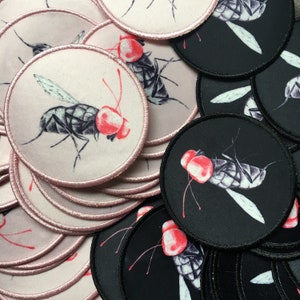 May include: A pile of round, fabric patches with a black and white illustration of a fly with red eyes. Some patches are pink and some are black.