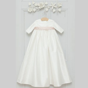 May include: A white satin christening gown with a pink satin sash. The gown has long sleeves and a round neckline.