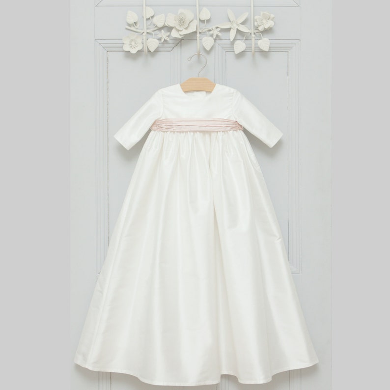 May include: A white satin christening gown with a pink satin sash. The gown has long sleeves and a round neckline.