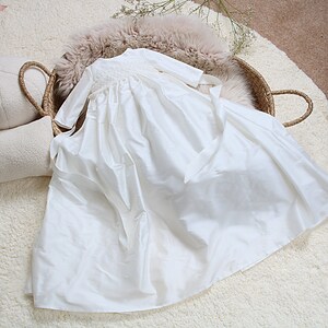 May include: White satin christening gown with long sleeves and a lace trim around the neckline. The gown has a sash tied around the waist.