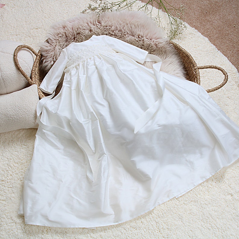 May include: White satin christening gown with long sleeves and a lace trim around the neckline. The gown has a sash tied around the waist.