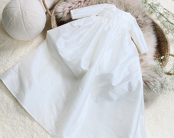Könnte beinhalten: Weißes Satin-Taufkleid mit langen Ärmeln und gerafftem Rock.
