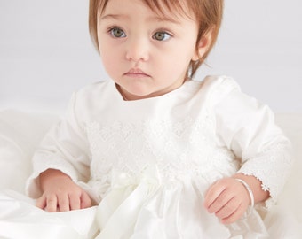 Könnte beinhalten: Ein kleines Mädchen in einem weißen Kleid mit Spitzendetails und einer weißen Schleife aus Satin. Sie trägt ein silbernes Armband am Handgelenk.