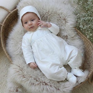 May include: A baby wearing a white outfit and bonnet lies in a woven basket on a fluffy, light-colored surface. The outfit includes a long-sleeved top and pants, with matching shoes. The baby is looking at the camera.