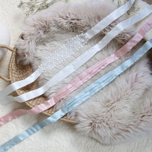 May include: Four satin ribbons in different colours: light blue, pink, white, and white with a lace trim. The ribbons are arranged on a fluffy white surface.