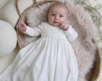 May include: A long-sleeved white christening gown featuring a pleated bodice and a full, flowing skirt. The garment is crafted from a smooth, solid white fabric with delicate horizontal stitching details across the chest area and long sleeves.