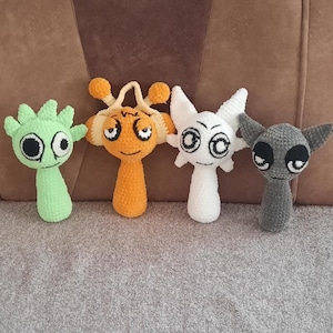 May include: Four crocheted plush toys in the shape of cartoon characters. The toys are green, orange, white, and gray. They have large eyes and are sitting on a brown couch.