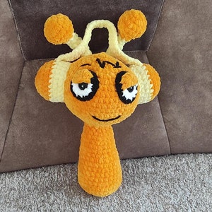May include: A crocheted orange and yellow toy with a face and two antennas. The toy has a grumpy expression with large black eyes.