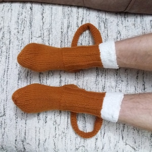 May include: A pair of knitted socks in a warm orange hue, featuring a white cuff and a loop handle. The socks are designed with a ribbed texture and are displayed on a light-colored surface. The socks are a cozy accessory.