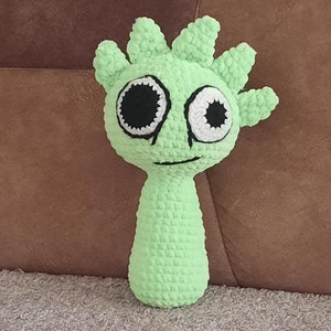 May include: A green crocheted toy with a spiky head and large white eyes with black pupils. The toy has a simple smile.