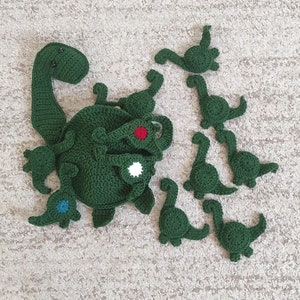 May include: A set of green crocheted dinosaur toys. The set includes a large dinosaur with a smaller dinosaur attached to its back, and several smaller dinosaurs that can be detached. The dinosaurs are all green and have black eyes.