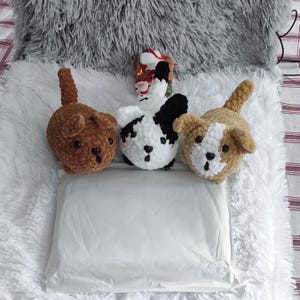 May include: Three crocheted stuffed animal dogs on a white fluffy surface. The dogs are brown, black and white, and brown and white. The dogs are all sitting up and looking at the camera.