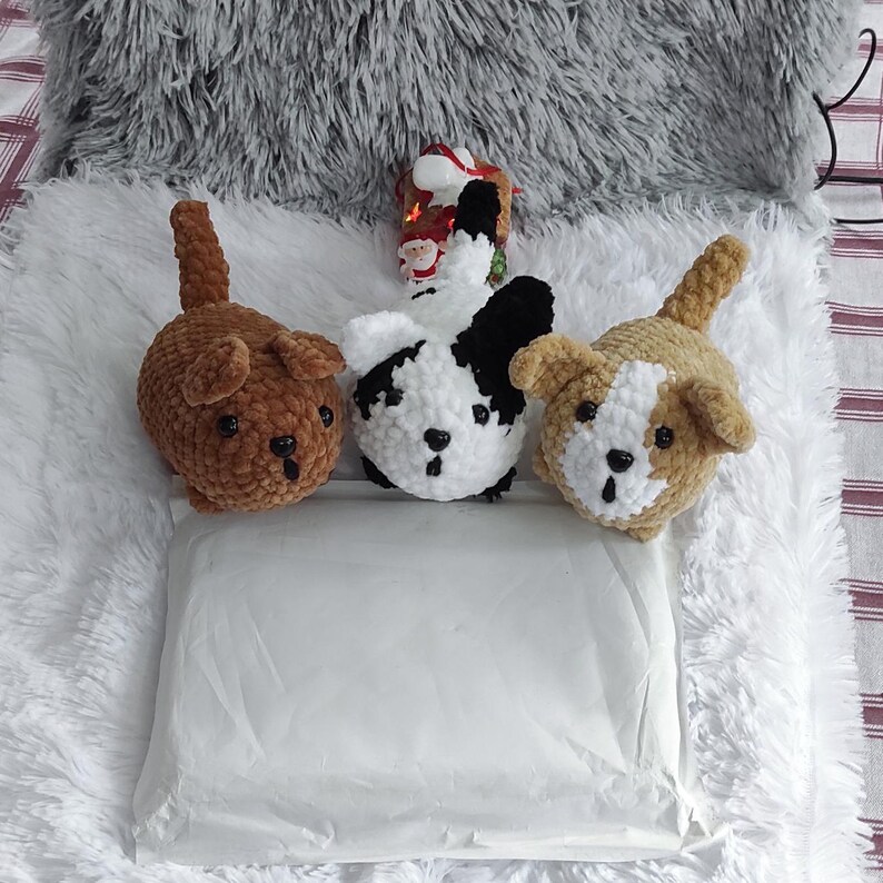 May include: Three crocheted stuffed animal dogs on a white fluffy surface. The dogs are brown, black and white, and brown and white. The dogs are all sitting up and looking at the camera.