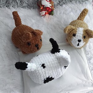 May include: Three crocheted stuffed animals shaped like dogs. One is brown, one is white with black spots, and one is tan with white accents.