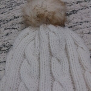 May include: White knitted beanie with a large, fluffy, cream-colored pom pom on top. The beanie has a cable knit pattern.