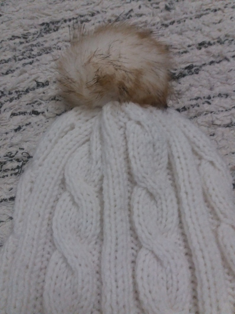 May include: White knitted beanie with a large, fluffy, cream-colored pom pom on top. The beanie has a cable knit pattern.