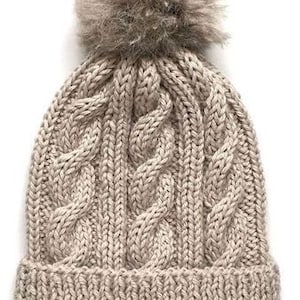 May include: A beige knitted beanie with a fluffy pom pom on top. The beanie has a cable knit pattern.