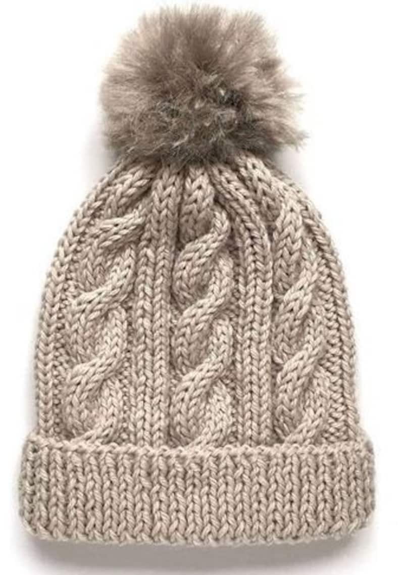 May include: A beige knitted beanie with a fluffy pom pom on top. The beanie has a cable knit pattern.