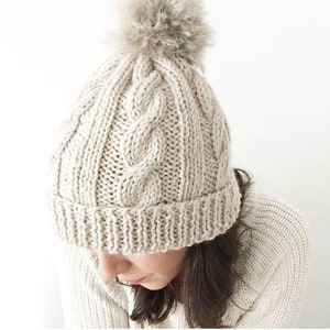 May include: A cream-colored knitted beanie with a cable knit pattern and a fluffy pom-pom on top.