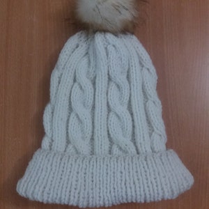 May include: White knitted beanie with a cable knit pattern and a fluffy pom pom on top.