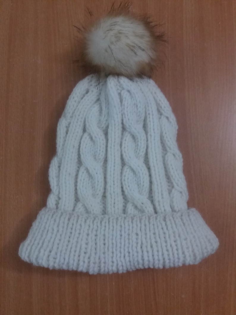 May include: White knitted beanie with a cable knit pattern and a fluffy pom pom on top.