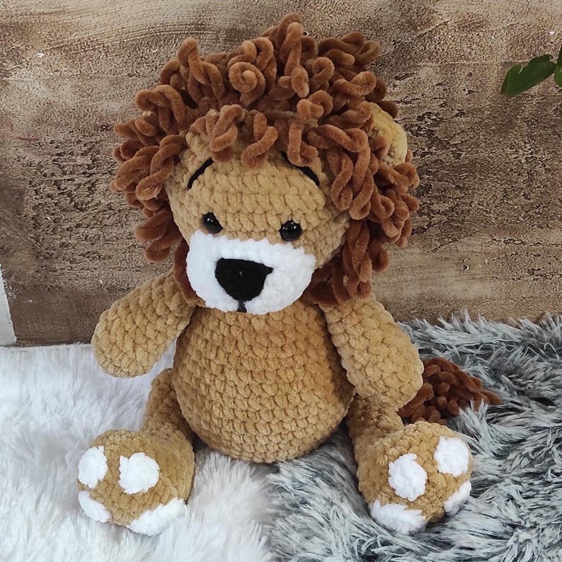 May include: A handmade plush lion toy with a tan body, brown mane, and white accents on the face and paws. The lion has black eyes and a black nose. The toy is sitting on a fluffy white and gray surface.