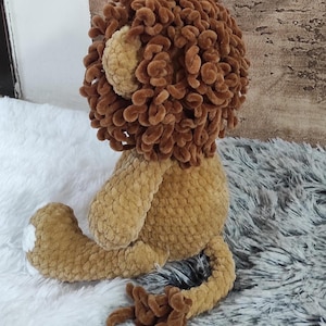 May include: A handmade crochet lion plush toy. The lion has a brown mane, tan body, and a white patch on its foot. The toy is sitting on a white and gray fluffy surface.