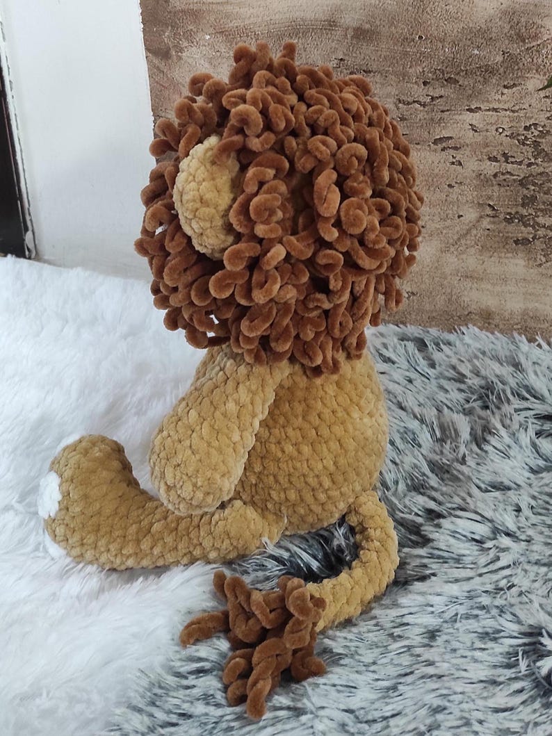 May include: A handmade crochet lion plush toy. The lion has a brown mane, tan body, and a white patch on its foot. The toy is sitting on a white and gray fluffy surface.