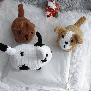 May include: Three crocheted stuffed animals in the shape of dogs. One is brown, one is white with black spots, and one is tan with white patches. They are all sitting on a white fluffy surface.