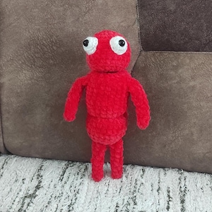 Pode incluir: Uma boneca vermelha de crochê feita à mão com olhos grandes brancos e pretos. A boneca tem uma cabeça arredondada, um corpo segmentado e braços e pernas simples. O brinquedo de pelúcia está em pé sobre um tapete branco, com um fundo marrom.