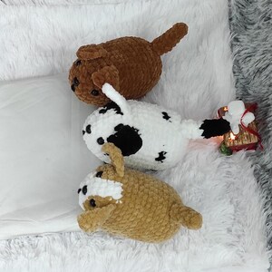 May include: Three crocheted stuffed dog toys. One is brown, one is white with black spots, and one is tan. The toys are lying on a white fluffy surface.