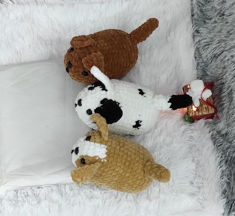 May include: Three crocheted stuffed dog toys. One is brown, one is white with black spots, and one is tan. The toys are lying on a white fluffy surface.