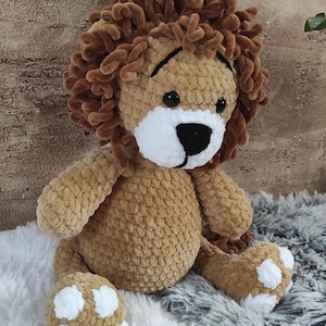 May include: A handmade, plush lion toy with a brown body and a fluffy, textured mane. The lion has a white muzzle, black eyes, and white paw pads. The toy is sitting on a soft, textured surface.