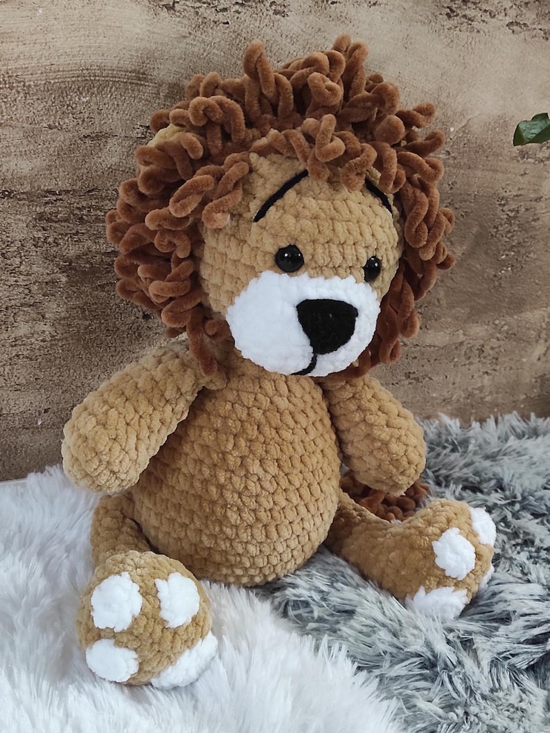 May include: A handmade, plush lion toy with a brown body and a fluffy, textured mane. The lion has a white muzzle, black eyes, and white paw pads. The toy is sitting on a soft, textured surface.