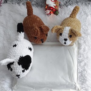 May include: Three crocheted stuffed dog toys. One is brown, one is white with black spots, and one is tan with white markings. The toys are sitting on a white surface.