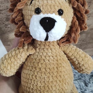 May include: A handmade plush lion toy with a tan body, brown mane, and a white muzzle. The lion has black eyes and is made of soft, textured yarn. A cuddly stuffed animal.