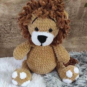 May include: A handmade, plush lion toy with a brown body and a fluffy, textured mane. The lion has a white muzzle, black eyes, and white paw pads with brown spots. The toy is sitting on a fluffy, textured surface.