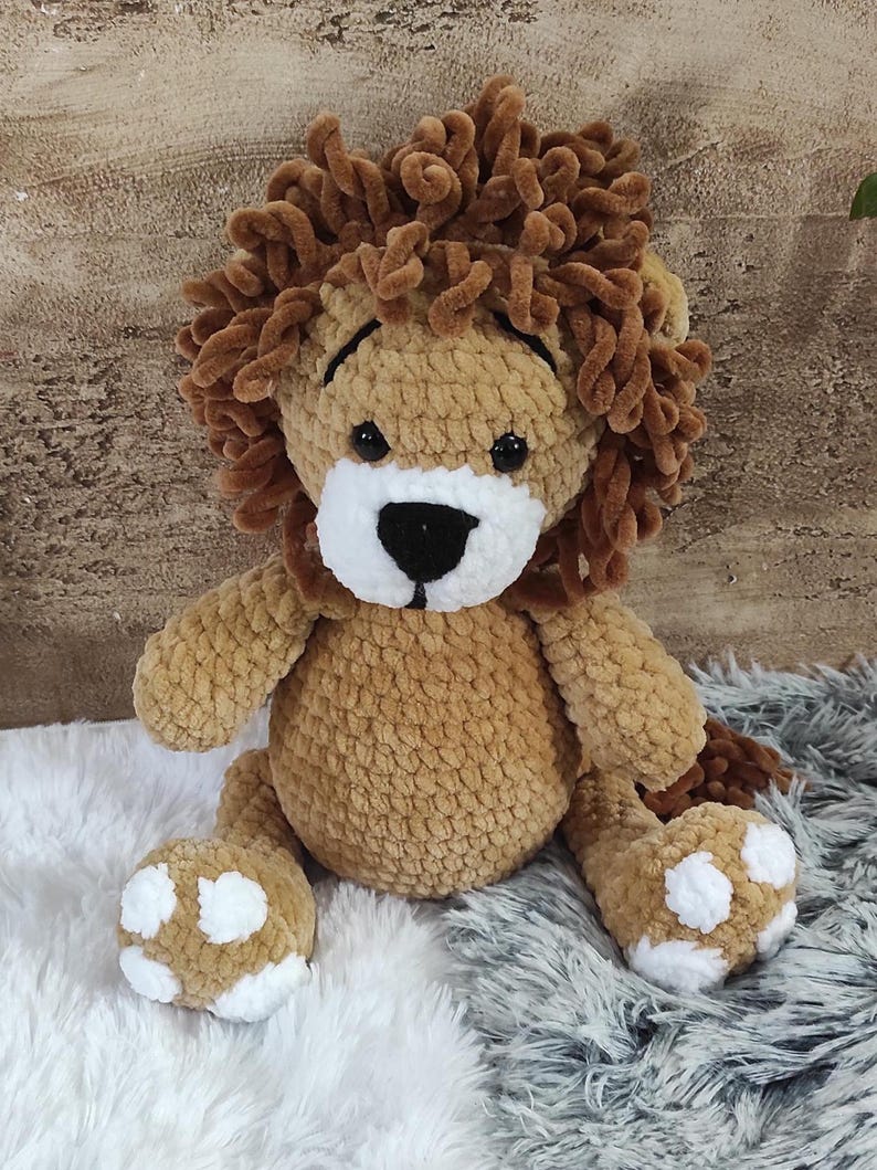 May include: A handmade, plush lion toy with a brown body and a fluffy, textured mane. The lion has a white muzzle, black eyes, and white paw pads with brown spots. The toy is sitting on a fluffy, textured surface.