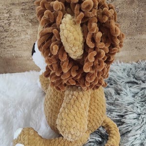 May include: A handmade plush lion toy in shades of brown and tan. The lion features a fluffy mane, ears, and tail, with white accents on the paws and face. The toy is made of soft, textured yarn, perfect for cuddling.