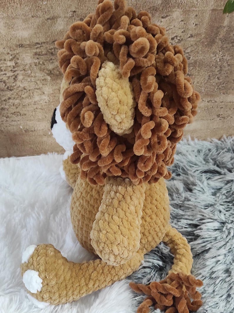 May include: A handmade plush lion toy in shades of brown and tan. The lion features a fluffy mane, ears, and tail, with white accents on the paws and face. The toy is made of soft, textured yarn, perfect for cuddling.