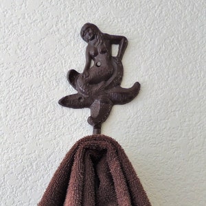 May include: A brown towel hanging on a cast iron mermaid hook. The hook is attached to a white wall.