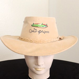 May include: A tan cowboy hat with a brown leather band and a green and white logo that says "The Coeur d'Alene" on the front.