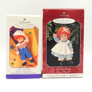 May include: Two Hallmark Keepsake ornaments featuring Madame Alexander dolls. One box is red with a doll in a blue dress, the other is white with a doll in a checkered shirt and blue pants. Text on the boxes reads "Mop Top Billy" and "Mop Top Wendy".