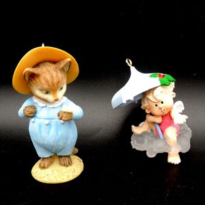 May include: Two small figurines on a black background. One is a cat wearing a yellow hat and blue outfit. The other is a cherub with wings, a red top, and a white umbrella, sitting on a cloud. Both are Christmas ornaments.